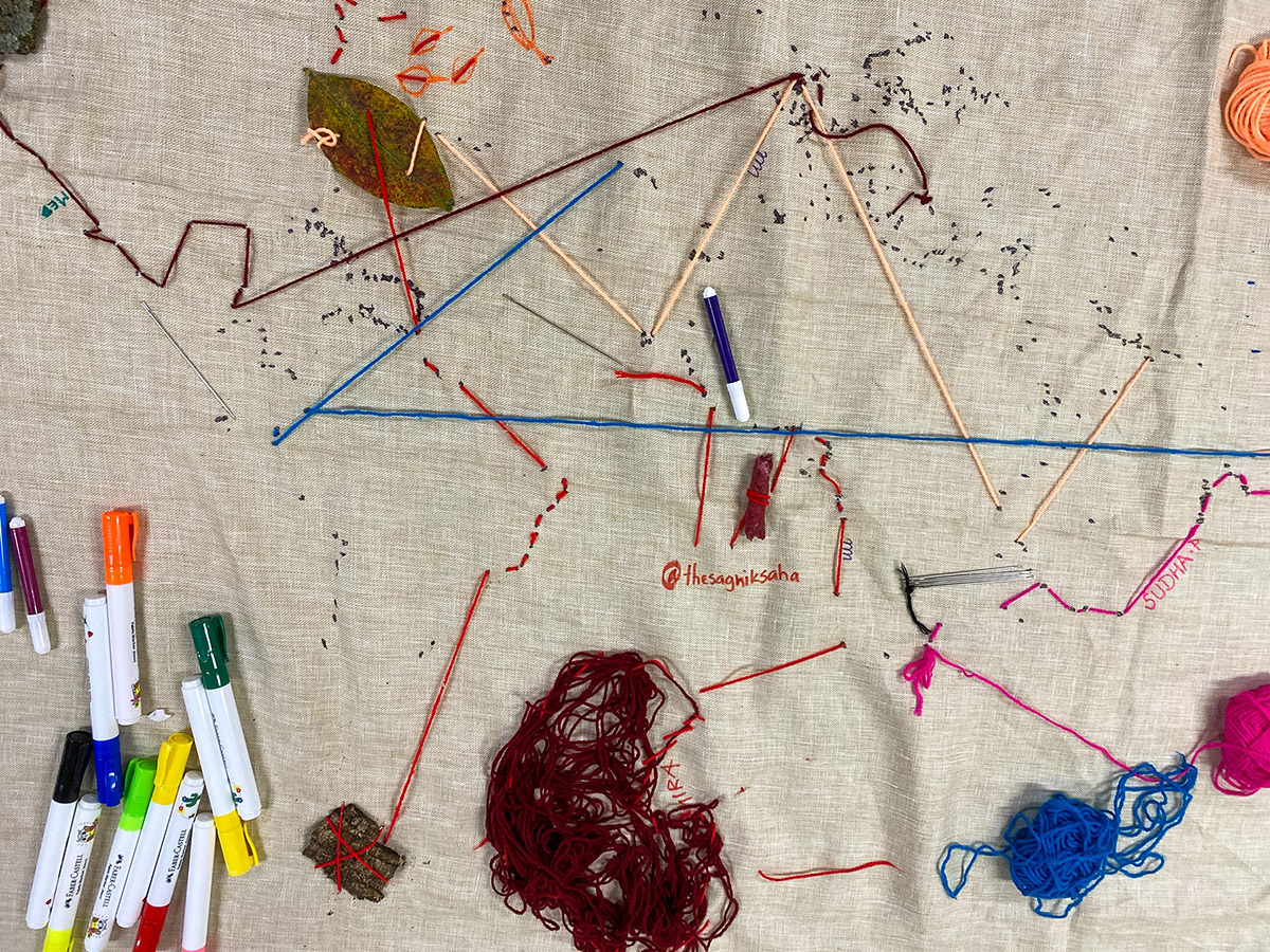 A large piece of grey-brown linen fabric is photographed from above. Black painted dots in the upper half of the fabric represent internet landing stations and participants have connected them in new ways by using yarn and natural materials. Strings in many colours enter and exit the fabric, forming an embroidered network. A leaf, pieces of bark, and knotted strings have been knitted into the fabric as well and form nodes. Across the fabric, pens, needles and balls of unused yarn are scattered.